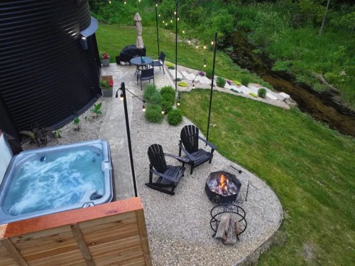 Aerial view of a backyard with a hot tub, fire pit, chairs, and string lights near a stream and lush greenery.