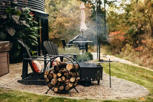 A cozy outdoor space with a fire pit, stacked firewood, and chairs surrounded by greenery and trees.