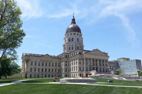A grand state capitol building with a dome, surrounded by green lawns and trees under a clear blue sky.
