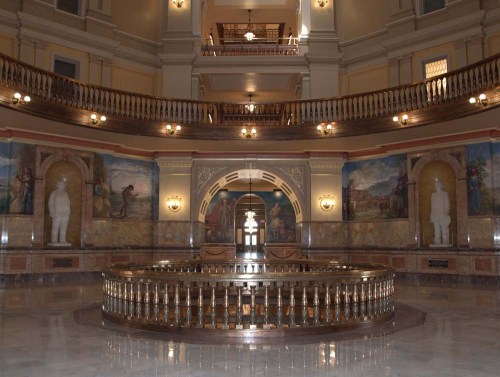 Interior view of a grand hall with ornate architecture, circular railing, and large murals on the walls.