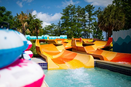 Colorful water slides twist and turn beside a pool, surrounded by trees and a bright blue sky.