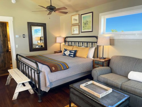 Cozy bedroom featuring a metal bed, gray sofa, wooden bench, and decorative artwork on the walls. Bright natural light.