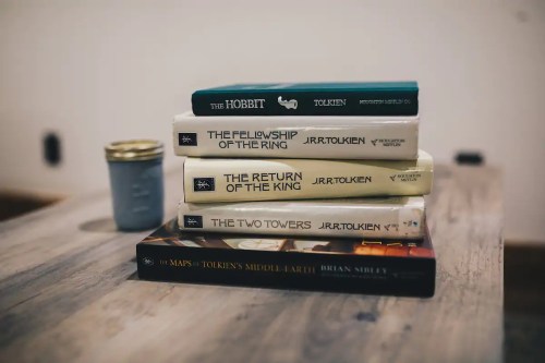 A stack of J.R.R. Tolkien books, including "The Hobbit" and "The Lord of the Rings," on a wooden table beside a jar.