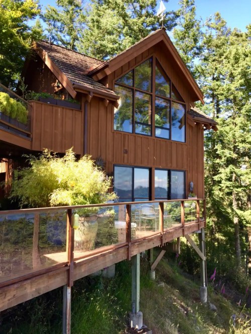A wooden house with large windows, surrounded by trees, featuring a deck with a glass railing.
