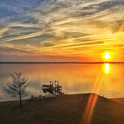 A serene sunset over a calm lake, with a dock and chairs silhouetted against vibrant orange and yellow skies.