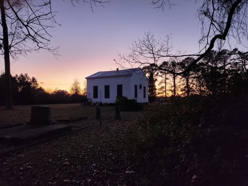 A white building stands in a field at sunset, surrounded by trees and gravestones, creating a serene atmosphere.