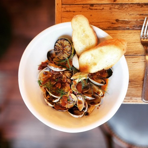 A bowl of clams in a savory sauce, garnished with herbs and served with two slices of toasted bread.
