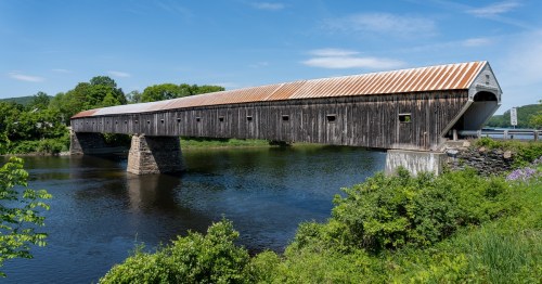 A wooden covered bridge spans a calm river, reflecting its structure in the water below, surrounded by greenery.