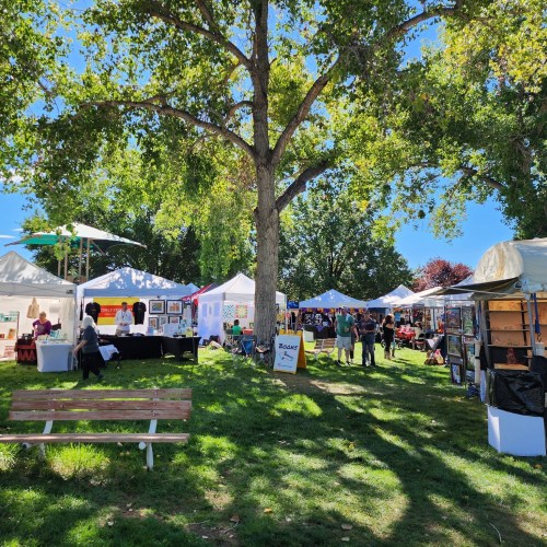 A vibrant outdoor market with tents displaying art and crafts, surrounded by trees and visitors enjoying the day.