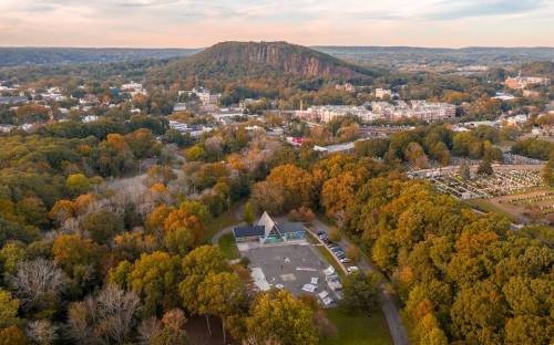 Aerial view of a town surrounded by autumn trees, featuring a triangular building and a hill in the background.