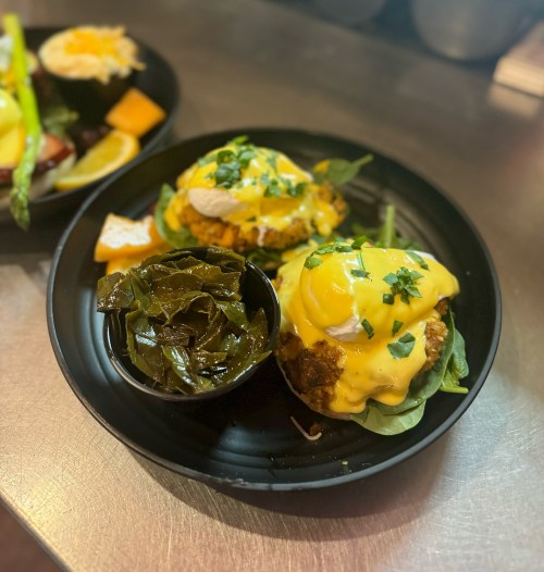 Two eggs Benedict on a black plate, topped with hollandaise sauce and green onions, served with a side of collard greens.