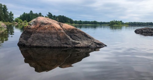 A large rock partially submerged in calm water, reflecting the surrounding trees and cloudy sky.