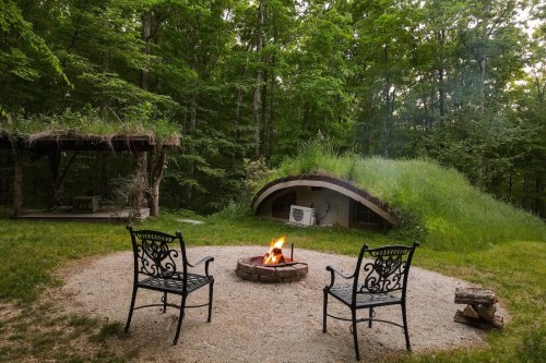 A cozy outdoor scene with two chairs around a fire pit, surrounded by trees and a unique grass-covered structure.