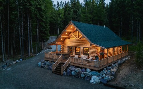 A cozy log cabin with a green roof, surrounded by trees, illuminated warmly at dusk. Stone pathway leads to the entrance.
