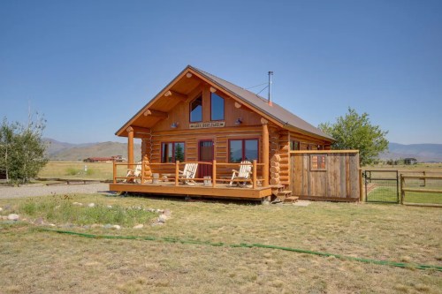 A cozy log cabin with a wooden porch, surrounded by open fields and mountains under a clear blue sky.