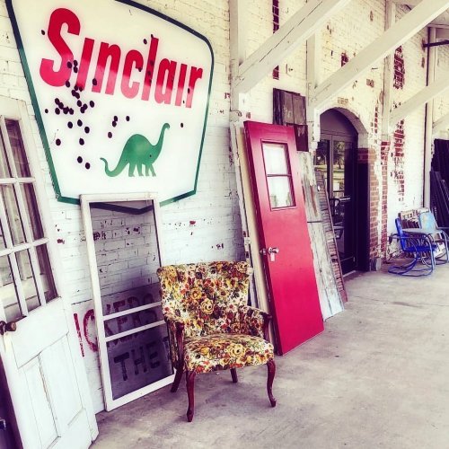 A vintage Sinclair sign with a dinosaur, a floral chair, and various doors in a rustic setting.