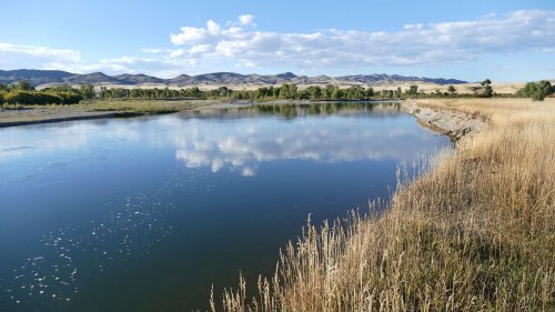 A serene river reflects clouds and mountains, surrounded by grassy banks and a clear blue sky.