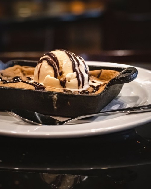 A warm chocolate chip cookie in a skillet topped with vanilla ice cream and drizzled with chocolate sauce.