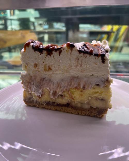 A slice of layered dessert with a graham cracker crust, banana, cream, and chocolate drizzle on top.