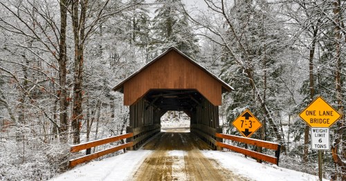 A rustic covered bridge surrounded by snow covered trees and foliage, spanning a small creek.
