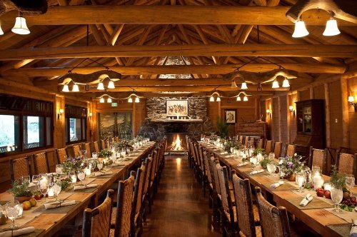 A rustic dining room with a long wooden table, elegant place settings, and a cozy fireplace, surrounded by log walls.