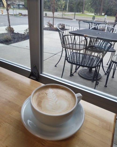 A cup of coffee with latte art sits on a wooden table, with outdoor seating visible through a window.