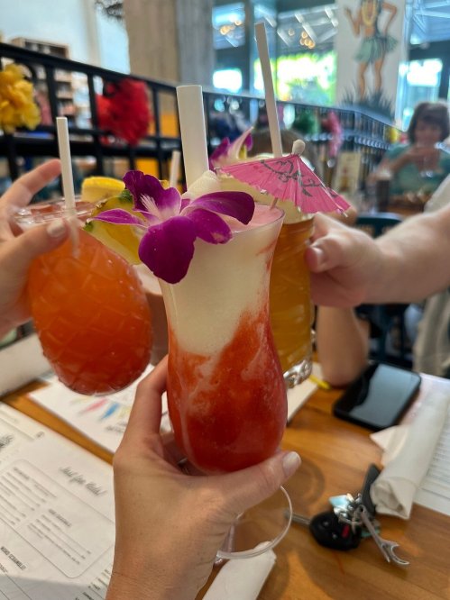 Three colorful cocktails are raised in a toast, featuring garnishes like flowers and umbrellas, in a lively restaurant setting.