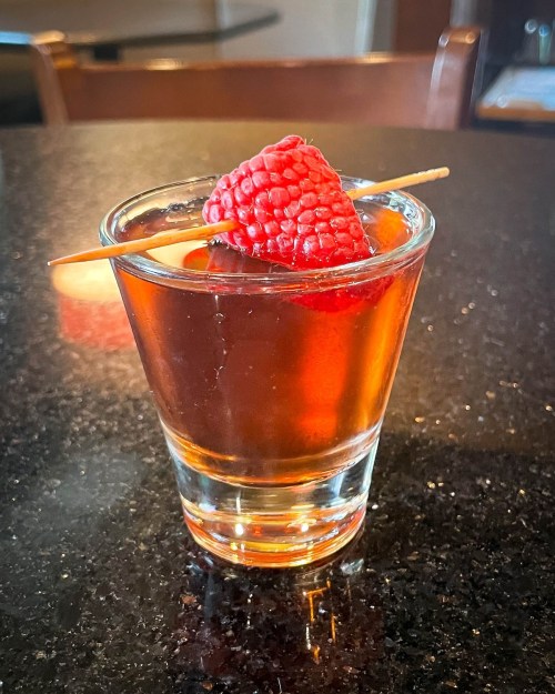 A glass of dark drink garnished with a raspberry on a skewer, resting on a black countertop.