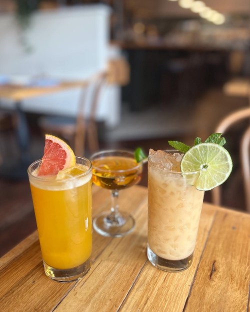 Three colorful cocktails on a wooden table: a yellow drink with grapefruit, a small glass of amber liquid, and a creamy drink with lime.