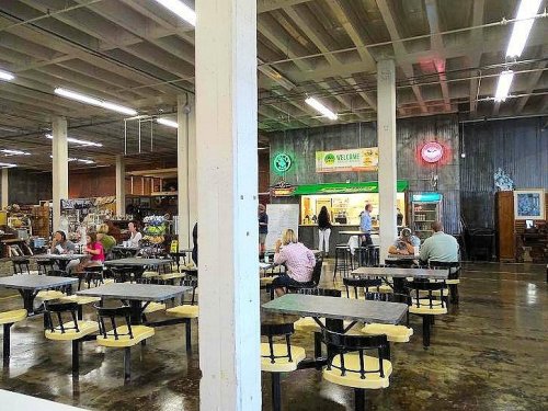 A spacious indoor market with tables, people dining, and various food stalls in the background.