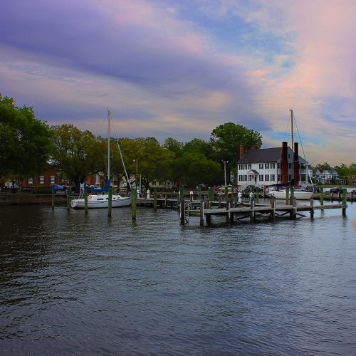 A serene waterfront scene with sailboats, a dock, and a charming house under a colorful sky.