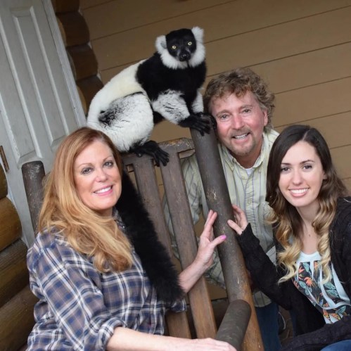 A woman, a man, and a girl pose together with a black-and-white lemur on a wooden railing.