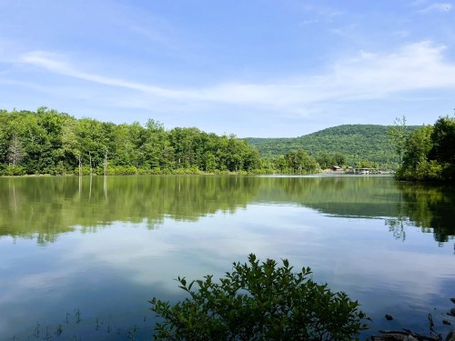 A serene lake surrounded by lush green trees and rolling hills under a clear blue sky. Reflections on the water.