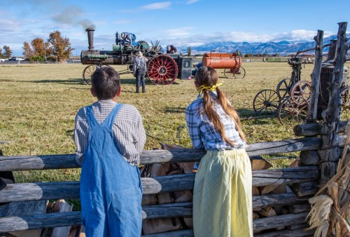 Two children watch a vintage steam engine at a rural event, with mountains in the background and autumn foliage.