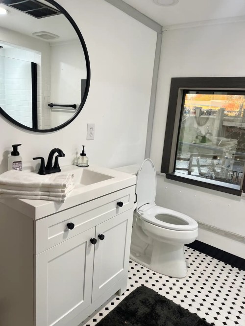 Modern bathroom featuring a round mirror, white sink cabinet, toilet, and black and white tiled floor.