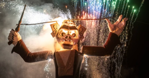 A large puppet figure with fireworks in the background, creating a festive and dramatic atmosphere at night.