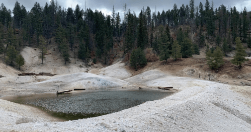 A serene landscape featuring a small pond surrounded by sandy hills and dense evergreen trees under a cloudy sky.