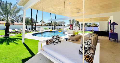 A modern outdoor space featuring a swing, pool, lounge chairs, and palm trees under a covered patio.
