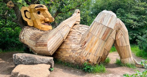 A large wooden sculpture of a reclining figure made from various pieces of wood, surrounded by greenery.