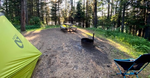 A serene campsite with a green tent, picnic table, fire pit, and a blue chair surrounded by trees and sunlight.