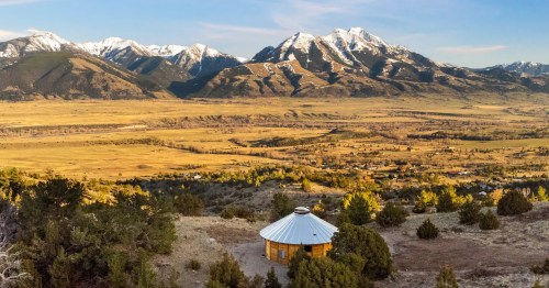 A scenic view of a round cabin in a valley, surrounded by mountains and a clear blue sky. Snow-capped peaks in the distance.