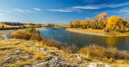 A serene river winds through a landscape of autumn trees and rocky banks under a clear blue sky.