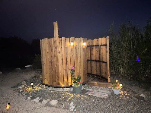 A wooden outdoor shower with lights, surrounded by plants and stones, set against a night sky.