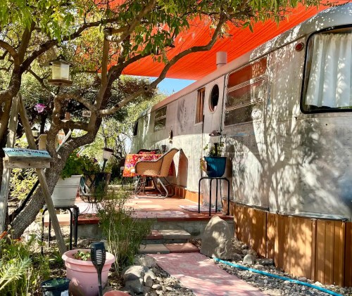 A vintage trailer surrounded by greenery, with a cozy seating area and vibrant orange awning overhead.