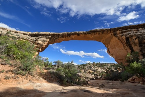Natural stone arch against a blue sky with scattered clouds, surrounded by desert vegetation and rocky terrain.