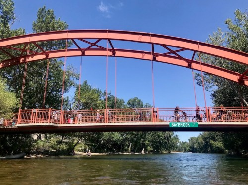 A red arch bridge spans a river, with people walking on it and trees lining the banks under a clear blue sky.