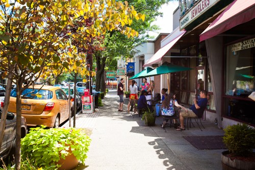 A sunny street scene with people sitting at outdoor tables, shops, and colorful trees lining the sidewalk.