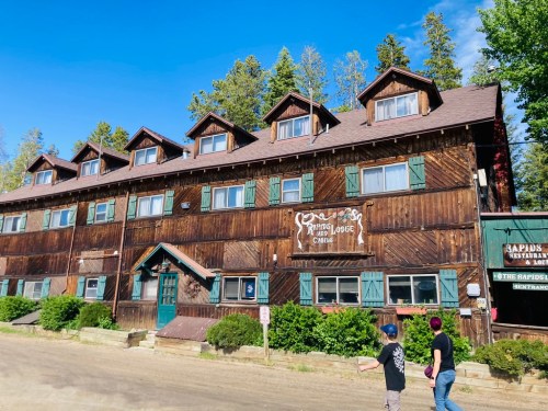 A rustic wooden lodge with multiple gabled windows, surrounded by trees and a dirt path in front.