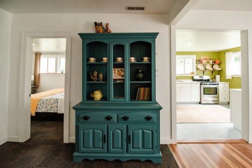 A teal cabinet with shelves displaying cups and decor, set in a cozy, light-filled room with a kitchen visible in the background.