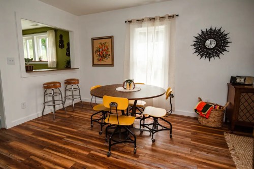 Cozy dining area with a round table, yellow chairs, and a window, featuring wooden floors and decorative wall art.